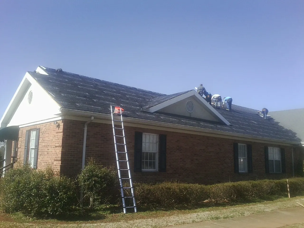 Crew tearing shingles off a brick ranch roof with a ladder set — Emergency Roof Repair work by our crew in Pinewood