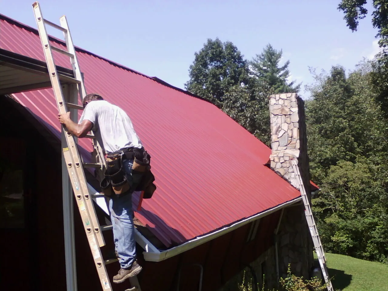 Roofer on a ladder beside a red metal roof and stone chimney — Soffit & Fascia Repair by Roofing Brothers in Pinewood